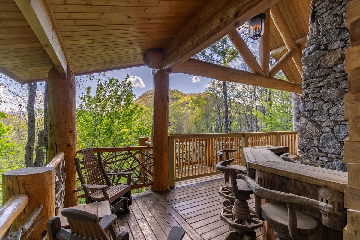 A spacious wooden deck with a stone wall and bar area, surrounded by trees and overlooking stunning mountain views. The setting invites groups to relax with drinks, enjoy social gatherings, and take in the scenic landscape.