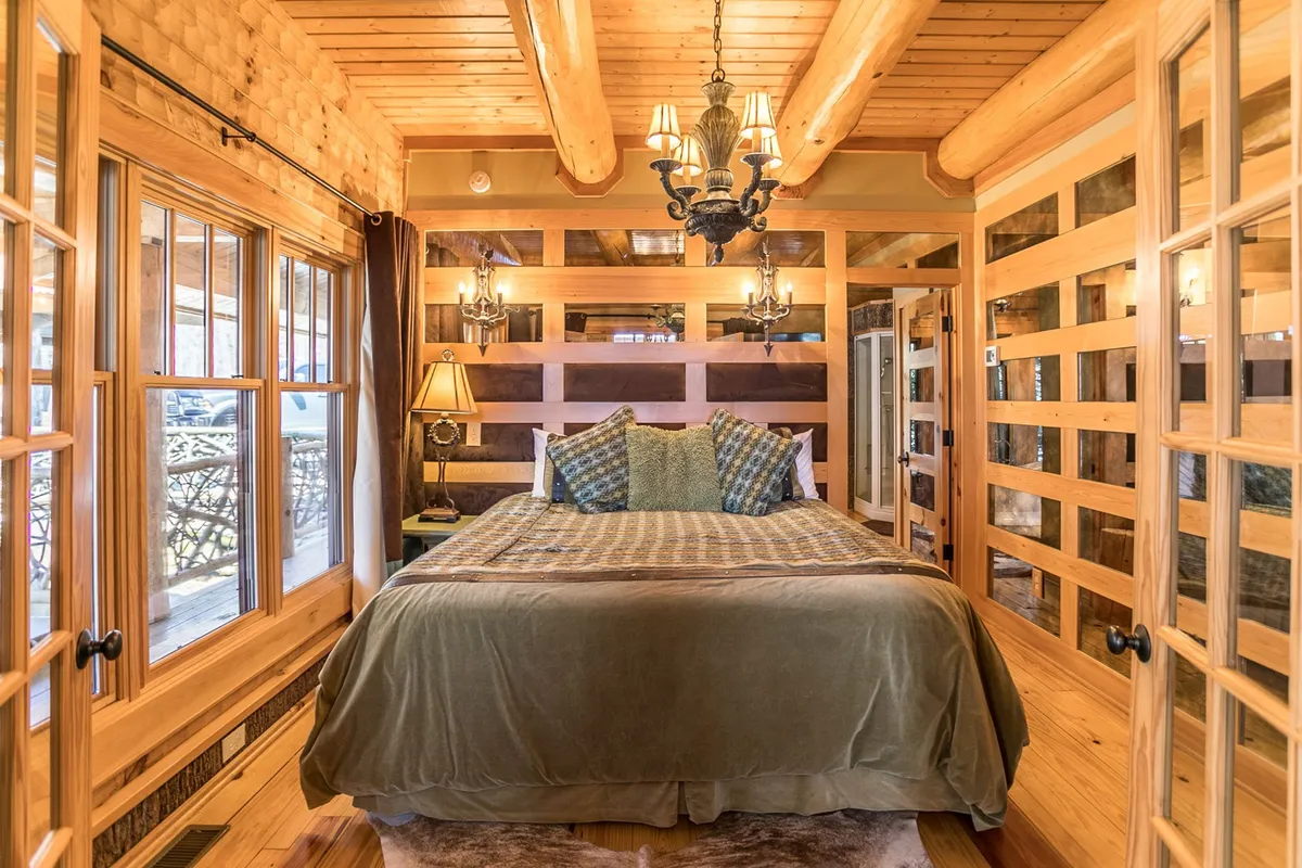 A tranquil bedroom featuring a large, plush bed with decorative pillows, rustic wooden beams overhead, a sparkling chandelier, and expansive windows that let in soft natural light. The room feels warm and restful, with cozy linens and elegant touches, inviting relaxation and peaceful sleep.