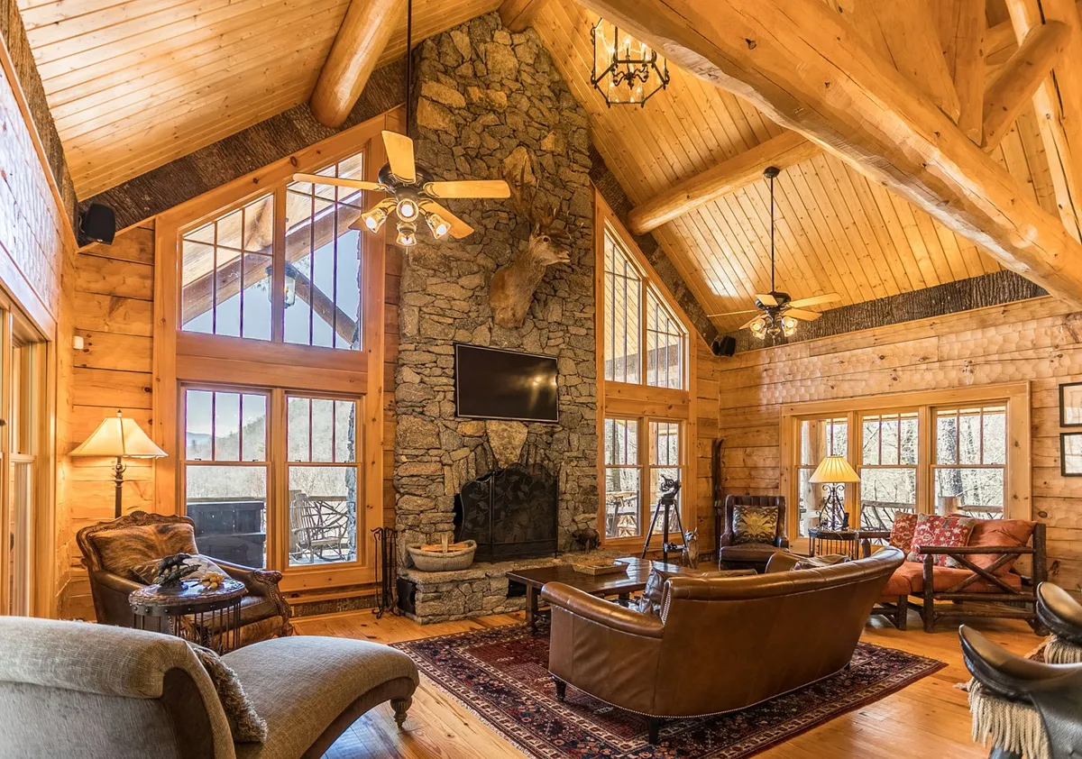 This interior image showcases a welcoming great room complete with a striking stone fireplace, expansive wooden beams, large windows framing the natural scenery, and plush leather sofas arranged for socializing. The rustic decor and abundant natural light create an inviting atmosphere that encourages gatherings and relaxation.