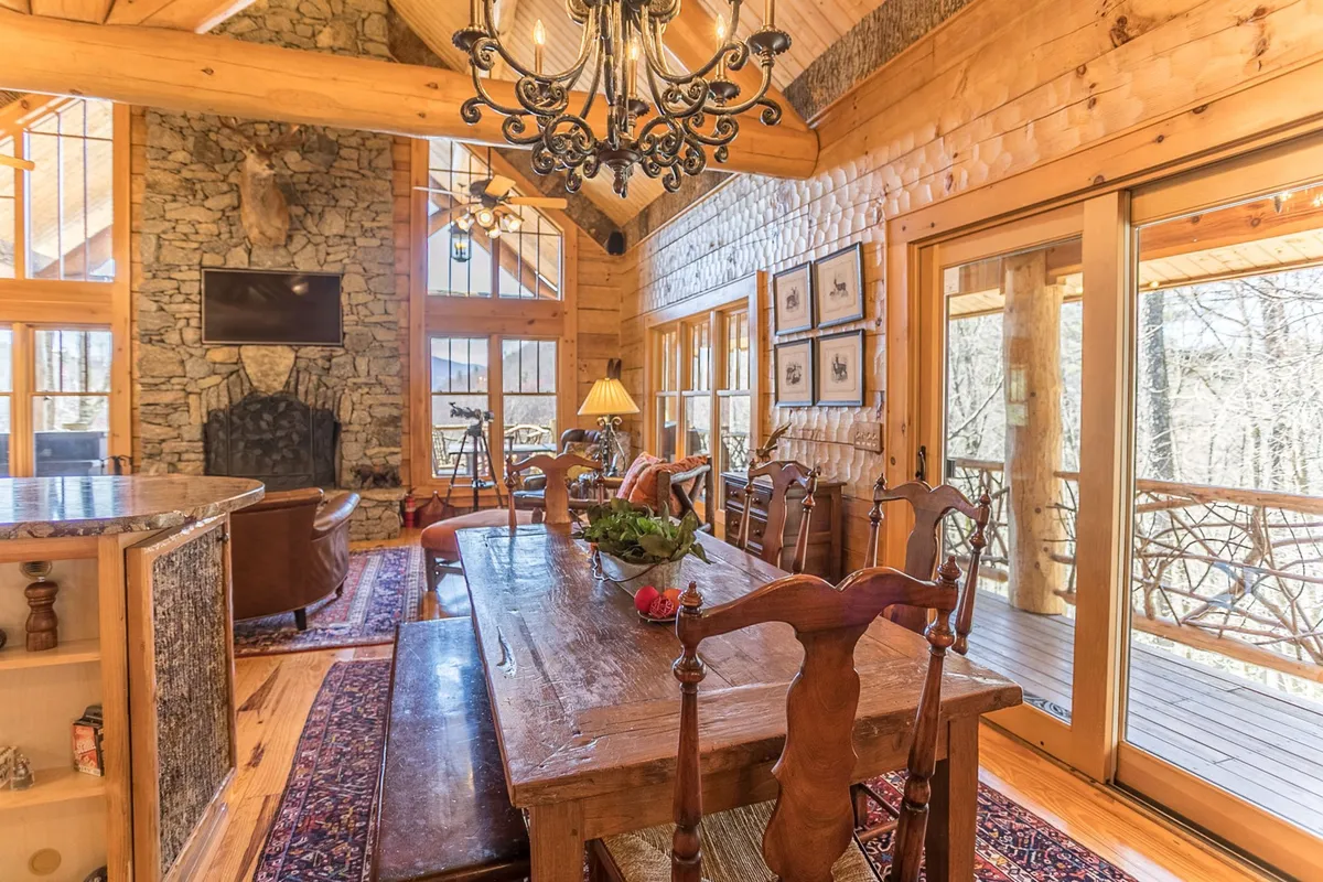 The spacious dining area features a large wooden table perfect for group meals, a cozy stone fireplace, elegant chandelier, and large windows offering scenic views. The natural wood beams overhead add to the rustic yet luxurious feel, ideal for lively group dinners or quiet mornings.