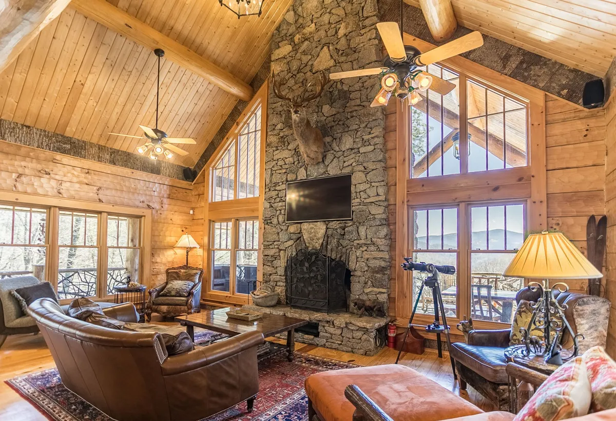 A cozy, upscale living room with a grand stone fireplace, leather sofas, wooden beams, and large windows framing tranquil natural views. The space invites relaxation by the fire, with a sense of warmth and comfort.