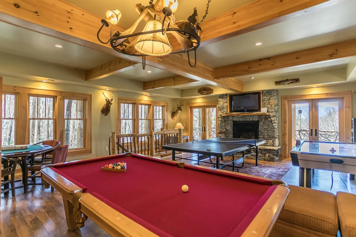 A lively recreation area with a pool table, ping pong table, and a cozy fireplace. Large windows provide scenic views and natural light, while a chandelier and rustic furnishings add warmth and character to the space.