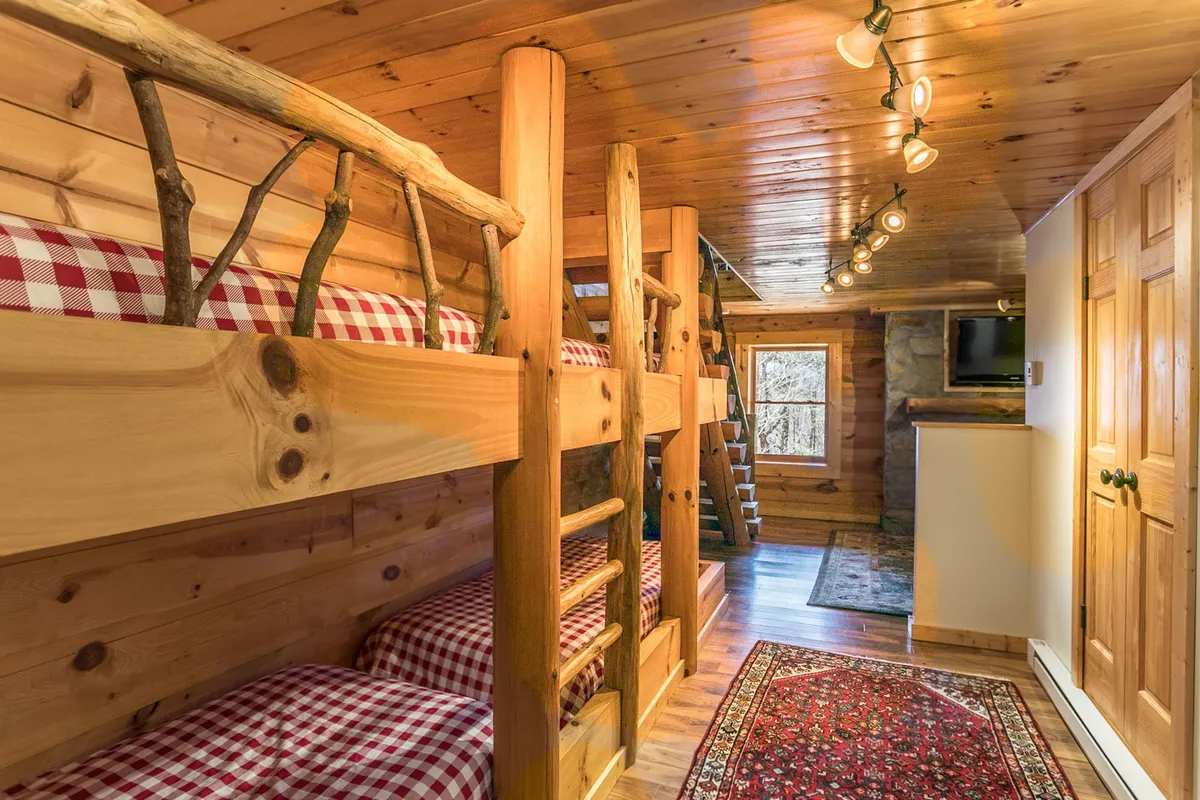 A cozy bunk room with sturdy wooden beams, inviting red checkered bedding on the bunk beds, and a soft rug on the floor. The warm lighting and playful arrangement suggest a perfect space for children, families, or groups to gather and rest.