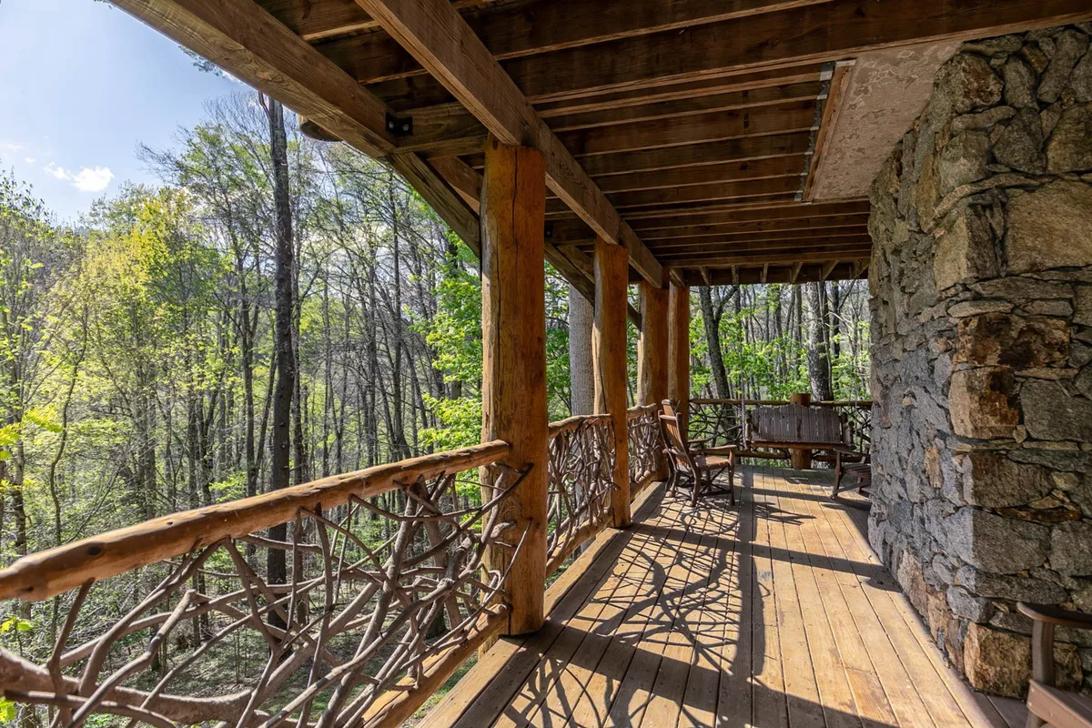 A tranquil porch scene featuring wooden railings, a stone wall, comfortable seating, and lush trees. The area is ideal for relaxing, reading, or enjoying coffee while immersed in nature.