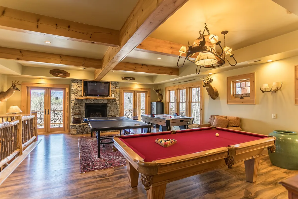 A spacious game room featuring a pool table at the center, a cozy fireplace in the background, game tables, and large windows letting in natural light. Comfortable seating is arranged for guests to relax between games. The rustic decor and open floor plan make it welcoming for all ages.