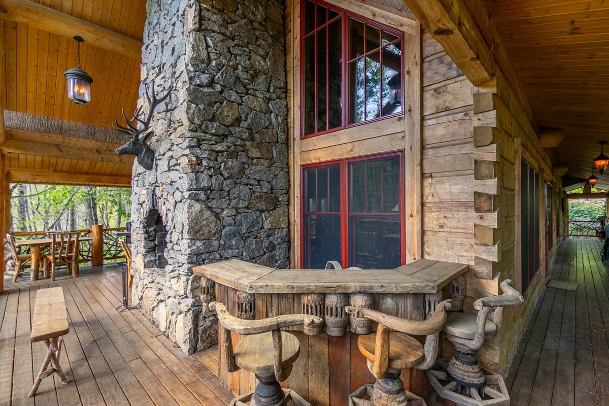 This image captures the covered deck area, featuring an inviting outdoor stone fireplace, rustic seating, a wooden bar, and open views to the natural surroundings. The scene is set for outdoor dining, fireside conversations, and enjoying the fresh mountain air.