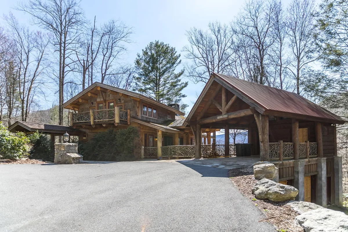 This image showcases the lodge's inviting wooden architecture, accented by stone features and surrounded by lush trees. The drive up to the home sets a tranquil, rustic tone, with visible porches and balcony spaces perfect for gatherings or relaxation. The natural setting offers a sense of peaceful seclusion and a direct connection to the outdoors.