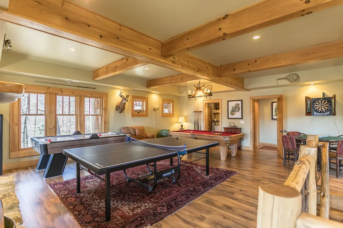 A comfortable and playful game room boasting both a ping pong and pool table, dartboard, wooden beams overhead, and a relaxed seating area for spectators or participants resting between matches.